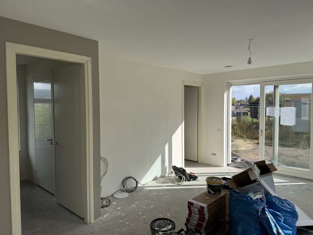 Pas opgeleverde woonkamer met kale wanden, binnendeuren, verfemmers en een schuifpui naar een bouwterrein