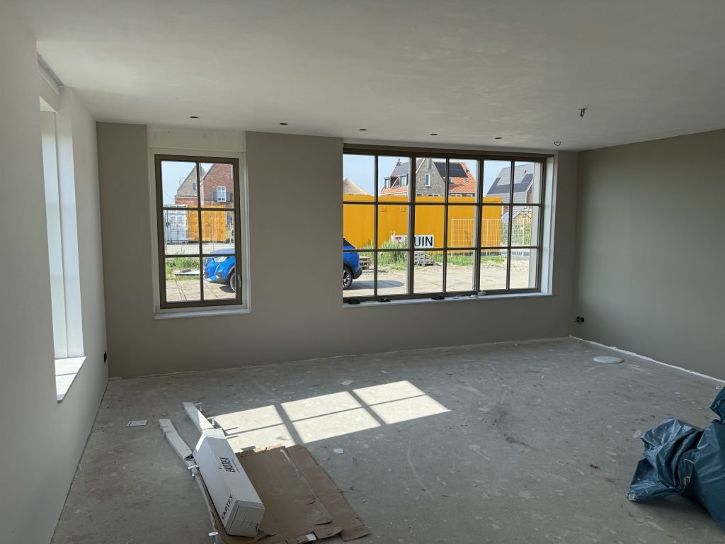 Nieuwbouwwoonkamer met strak afgewerkte grijze wanden, een betonvloer en ramen met roedeverdeling naar de straat