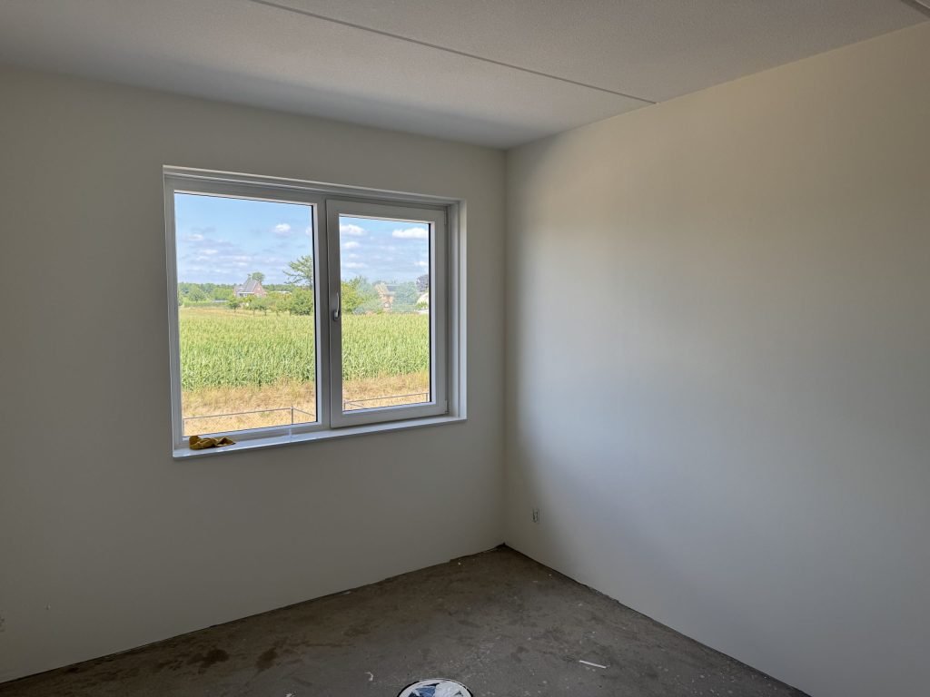 Kale nieuwbouwslaapkamer met gladde witte wanden en een raam met uitzicht op een maisveld