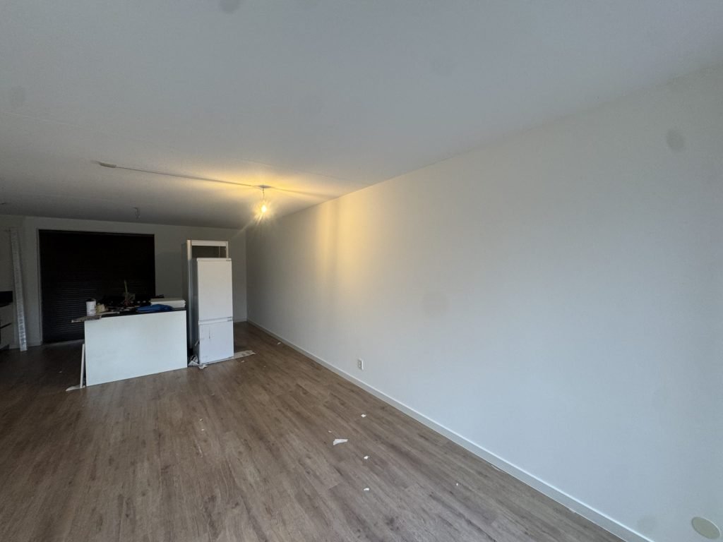 Lange lichte wand in een net opgeleverd appartement met open keuken, laminaatvloer en een brandende lamp aan het plafond