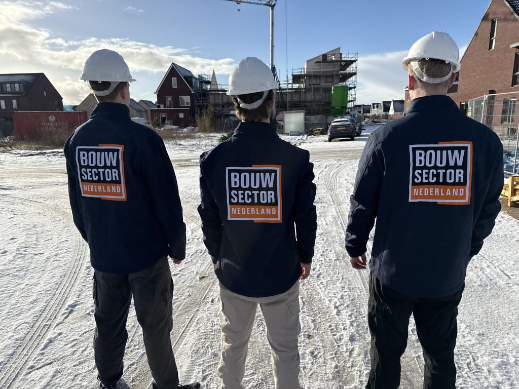 Drie vakmensen van Bouwsector Nederland met witte helm en jas met logo op een besneeuwde bouwplaats met nieuwe woningen en een bouwkraan